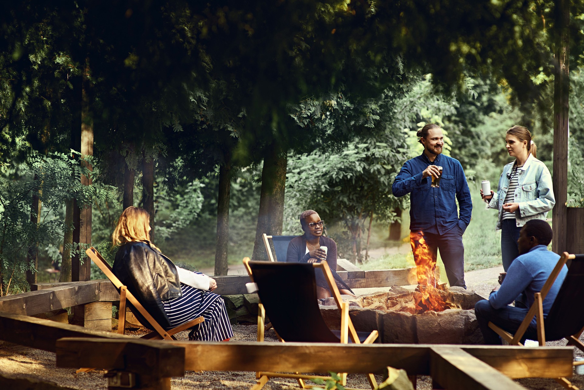 Multicultural group of friends enjoying beer by the campfire on a summer evening. Camping, tourism, holiday, outdoors concept. Barbeque party. Corporate team building event. Active lifestyle.